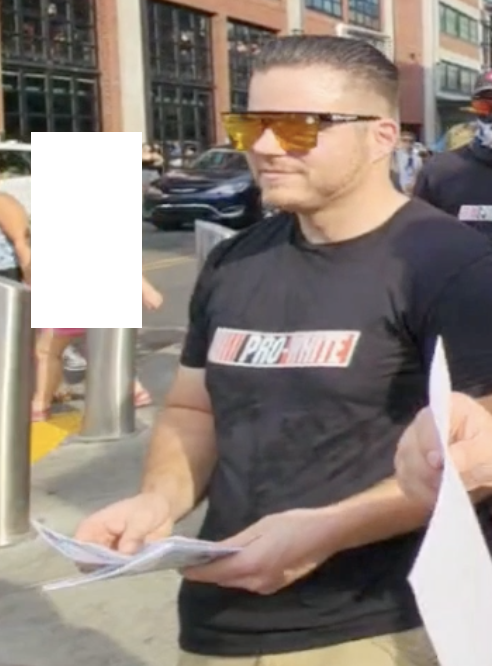 Colby Frank stands on a sidewalk in the daytime holding flyers. his black t-shirt says PRO-WHITE in NASCAR font. he wears his GDL totenkopf sunglasses.