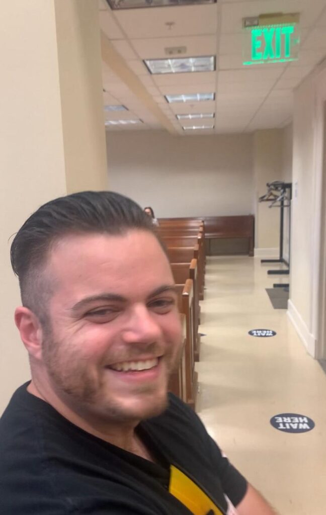 Colby Frank laughs at something off camera. he is sitting in an empty hallway outside a city council meeting his group has just disrupted.