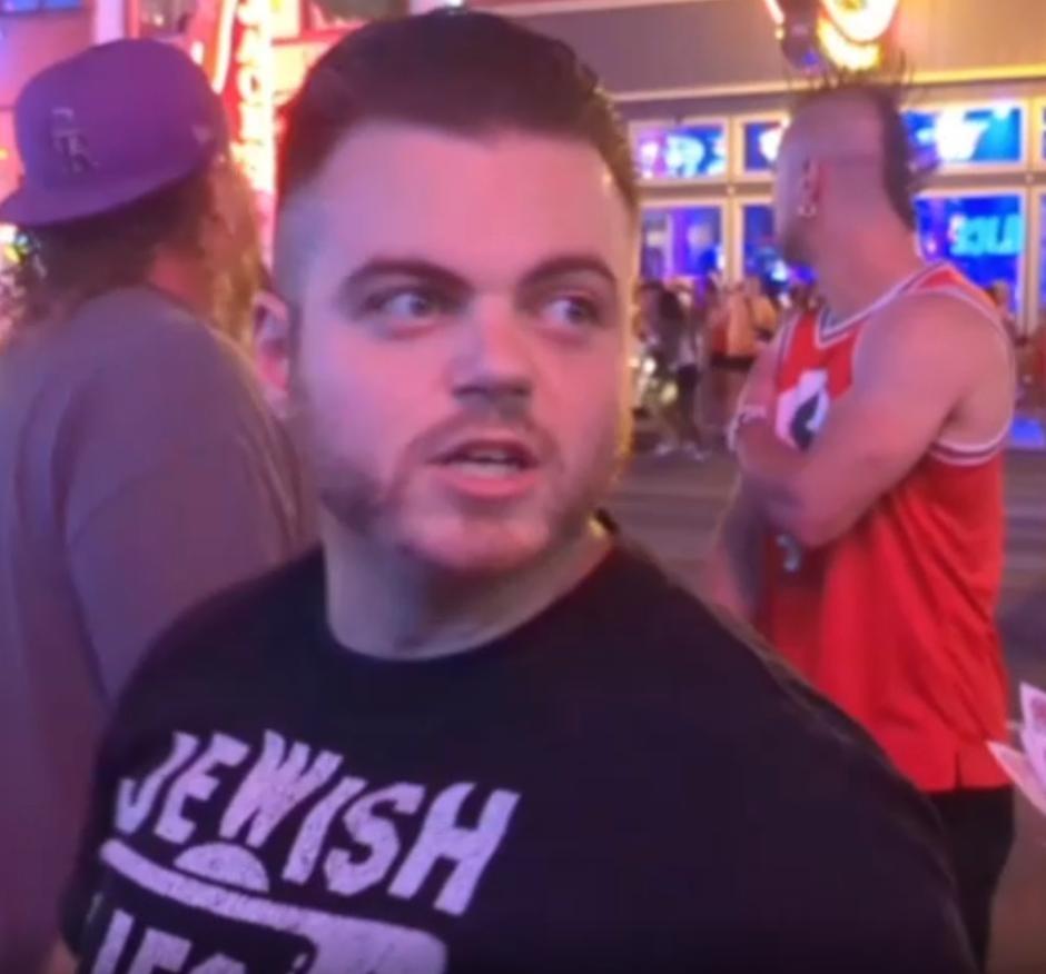 a 30ish white man with dark hair faces the camera, a busy street at night in the background. his black t-shirt has the word JEWISH visible in white at the top; the rest is cut off. this is Colby Alexander Frank. two men in the near background face away from the camera; one is wearing a red shirt with a white circle and part of a black swastika visible in the center, the Nazi flag.