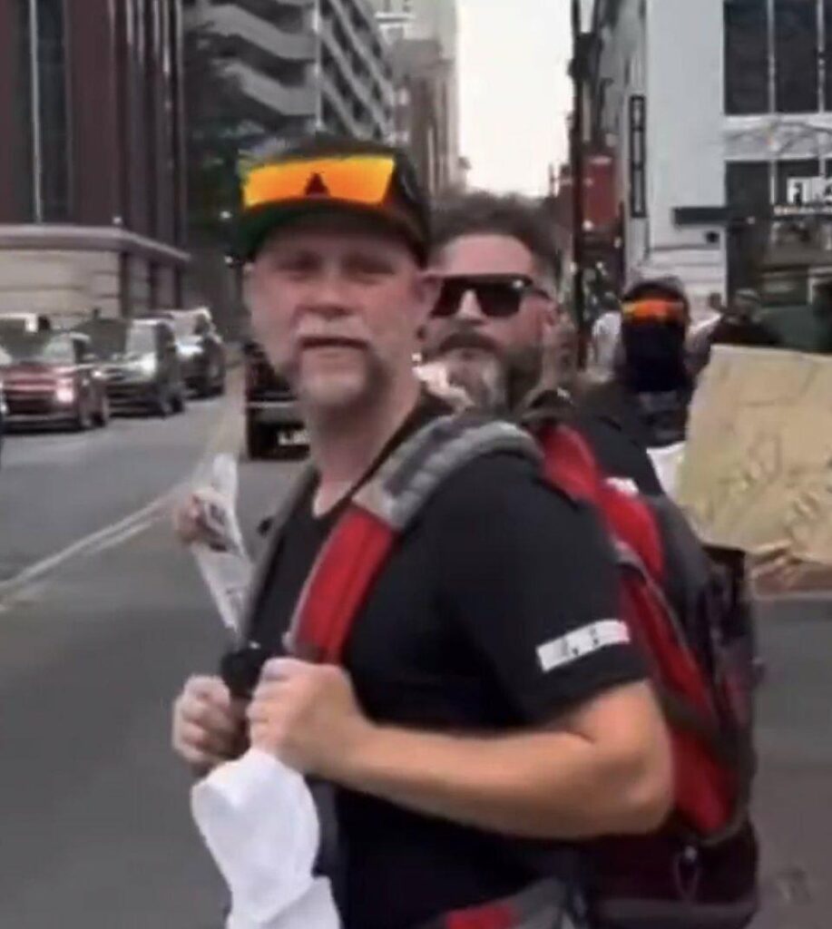 We see Jeffrey Perrine, a middle aged white guy with a blonde and patchy grey beard and a baseball cap with reflective sunglasses propped up on the brim. He clings to his backpack straps while walking down a city street next to a bearded man in sunglasses who is GDL leader John Minadeo aka Handsome Truth.