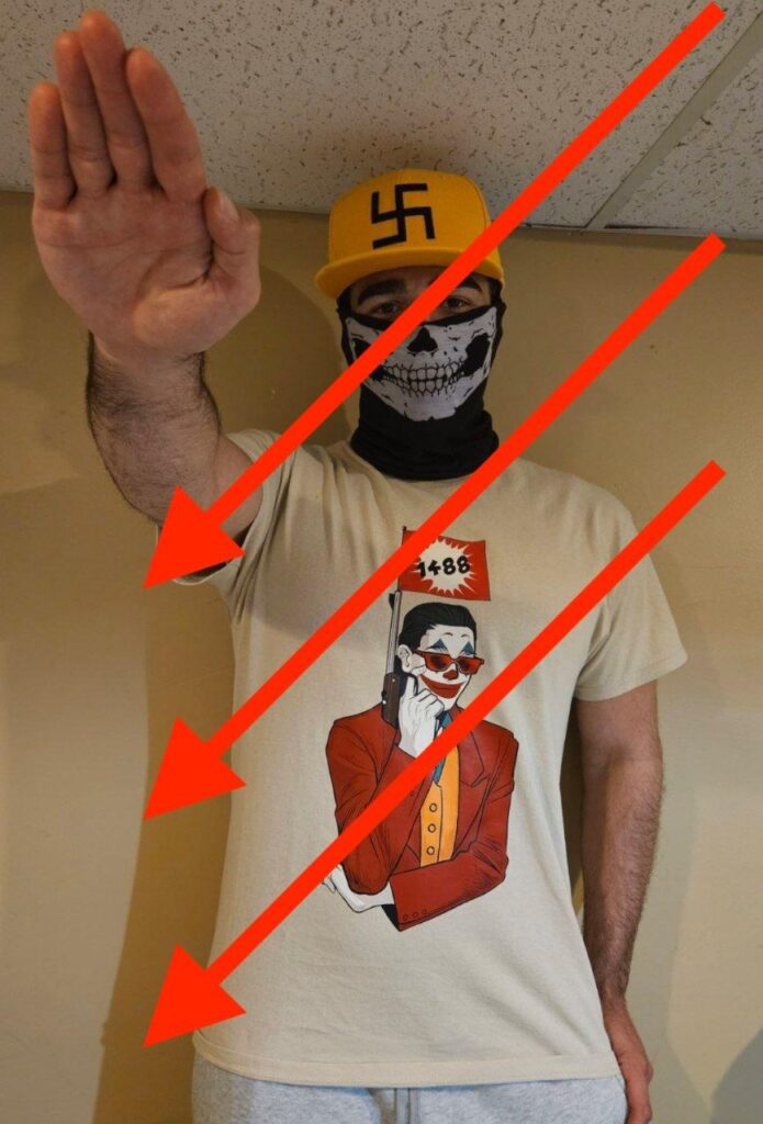  man gives a Nazi salute to the camera. He is standing indoors and wearing a yellow flat-brimmed hat with a swastika on it, a skull mask, and a shirt fraturing the image of a man wearing a Joker costume, holding a gun with a flag coming out of the end. The flag has the number “1488” on it. 