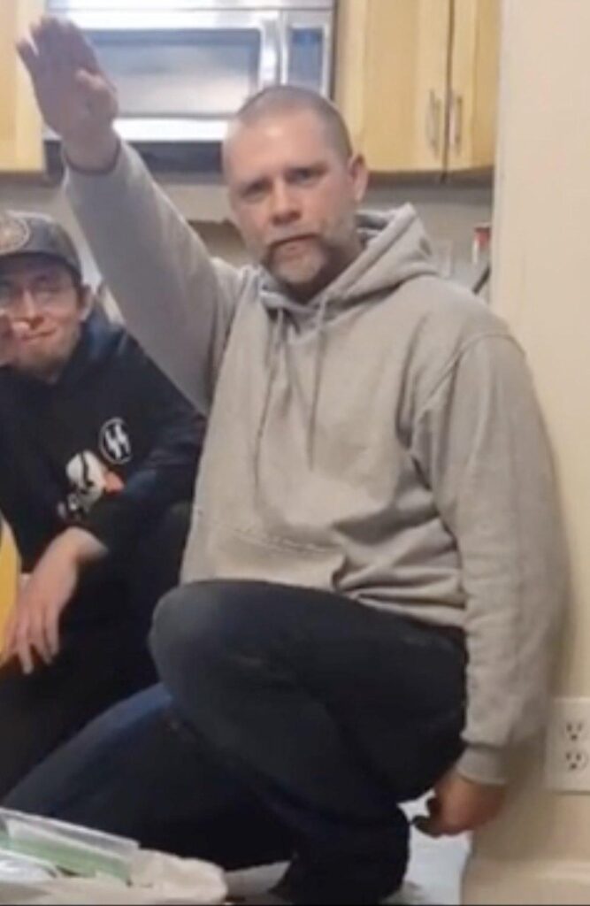 Jeffrey Perrine, a middle-aged white man with a shaved head and short, graying beard, squats in a kitchen, holding his arm up in a stiff-armed nazi salute. behind him is Harley Petero, another neo nazi, wearing a hat with a sonnenrad and the dual sig runes of the SS on his hoodie. 