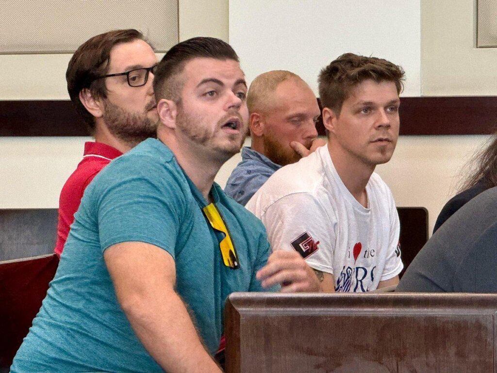 four men sit on a bench in a courtroom. Colby Frank is closest to the camera, wearing a turquoise shirt with GDL totenkopf (Nazi skull) sunglasses hanging from the neck. his expression is surprised. two of the other men have been identified in the #NameTheBozoTour as Nicholas Bysheim and Antoni Knezovic.