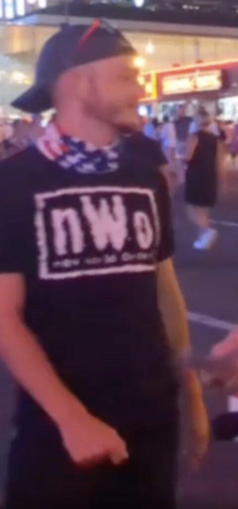 a blurry still from a video showing a young white man wearing a dark baseball cap backward, an American flag patterned cloth around his neck, a black nWo shirt, and black shorts or pants. he is in profile and a light beard is visible as well as glasses or sunglasses propped over his hat above his forehead. the background shows a city street at night with people walking by.