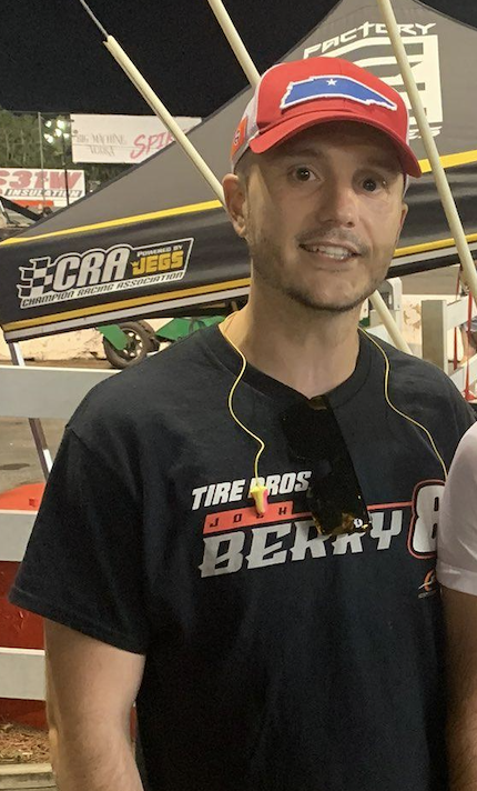 photo of a young white man, half-smiling-- a wider view of one of the photos from the first tweet. the man has a pair of distinctive flat rimless square-looking sunglasses hanging from his black t-shirt. a small white stylized skull logo is visible in the corner of one lens.